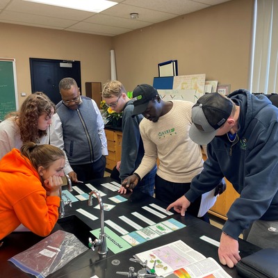A group of people gathered around a desk with informational papers spread out. A man is actively explaining something while the others listen attentively.