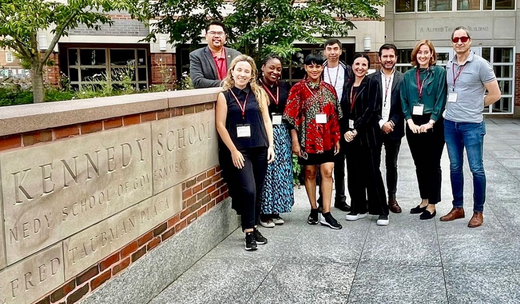 Individuals from Harvard University, both men and women, gathering for a group photo beside the Kennedy School wall.