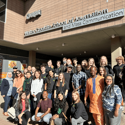 A gathering of individuals smiling for a photo in front of the School of Journalism building.