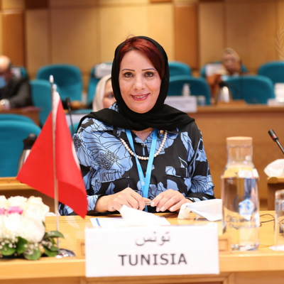 A smiling woman with a scarf around her head, seated at a desk in a hall with chairs.