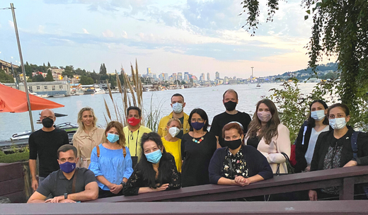 Individuals wearing masks assembled for a photograph, framed by a picturesque backdrop of a lake and distant buildings at the University of Washington.